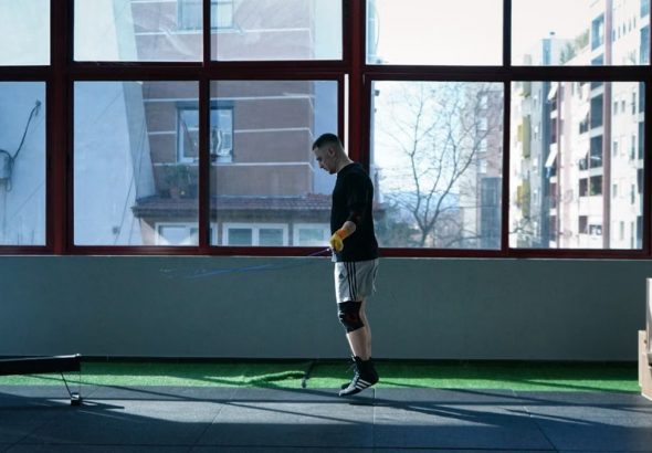 Young man exercising with a skipping rope in an urban gym with large windows and city view.